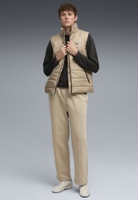 Beige puffer vest med høj krave, båret over en sort langærmet skjorte. Beige sweatpants og lyse sko fuldender outfittet.