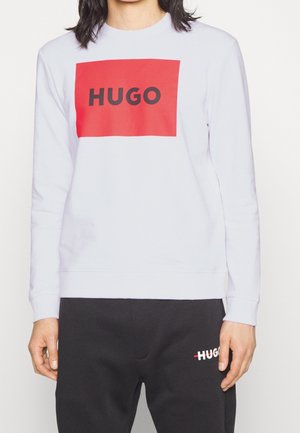 Persoon draagt een witte trui met een grote rode vierkant en zwarte tekst "HUGO", gecombineerd met een zwarte broek met klein "HUGO" logo.