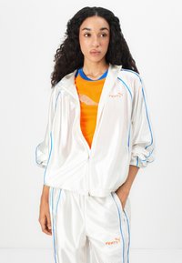 Tuta di satin bianco con accenti azzurri e logo FENTY. Sotto, una maglietta arancione con rifiniture blu, caratterizzata da una grafica del logo argentato.