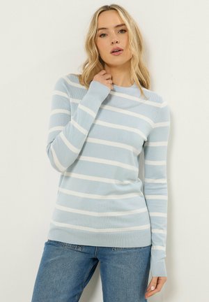 Vaaleahiuksinen nainen, joka on pukeutunut vaaleansinisillä ja valkoisilla raidoilla varustettuun pitkähihainen villapaitaan ja sinisiin farkkuihin, seisoo yksinkertaista valkoista seinää vasten.