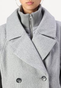 Manteau gris à double boutonnage avec un tissu texturé, de larges revers et un col côtelé haut avec une fermeture éclair à l'avant. Deux boutons noirs soulignent le devant.