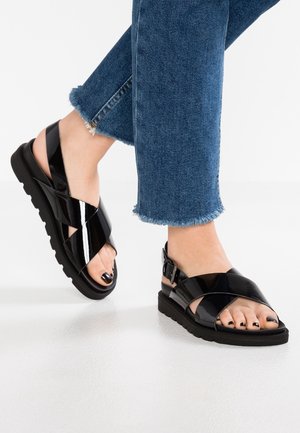 Sorte lakerede sandaler med krydsgående stropdesign og justerbar ankelspænde, med gummisål og åben tå.