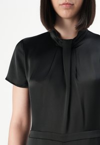 Woman wearing a black satin short-sleeve blouse with a high collar and pleated detail at the neckline against a white background.