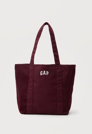 Donkerrode canvas tas met twee lange handvatten en witte "GAP" letters gedrukt nabij het bovenste midden.