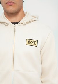 Felpa zip in off-white con finitura testurizzata. Presenta una toppa del logo EA7 Emporio Armani in nero e oro sul lato sinistro del petto.