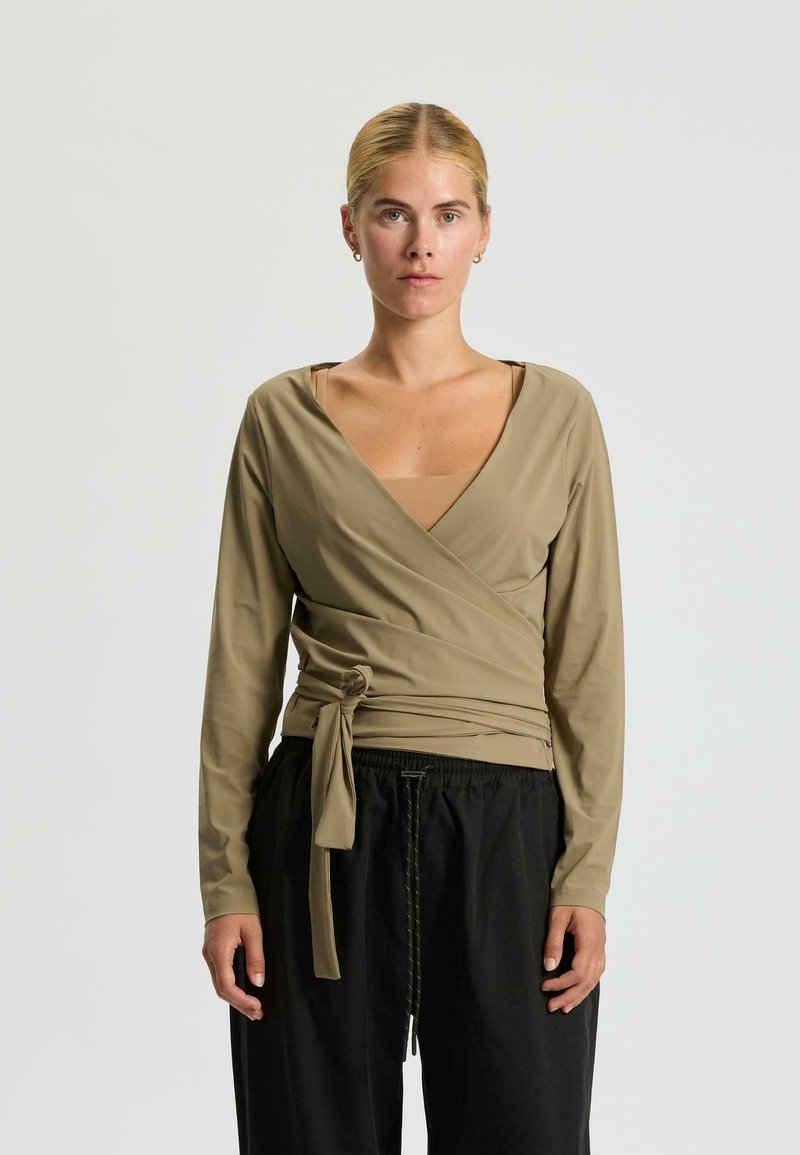 Haut enveloppé en tissu beige avec des manches longues, un décolleté en V profond et une lien à la taille, associé à un pantalon noir ample.