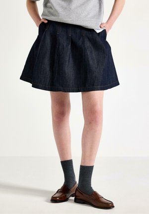 Persona che indossa una gonna plissettata in denim scuro, calzini grigi, mocassini marroni e una maglietta grigia, in piedi con le mani nelle tasche su uno sfondo semplice.