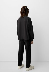 Osoba s dredovima nosi crnu majicu dugih rukava, crne traperice i bijele tenisice, stoji okrenuta prema praznom bijelom zidu.