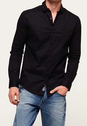 Mann trägt ein eng anliegendes schwarzes, langärmliges Hemd mit Kragen und Knöpfen sowie blaue Jeans, eine Hand in der Tasche, vor einfarbigem Hintergrund.