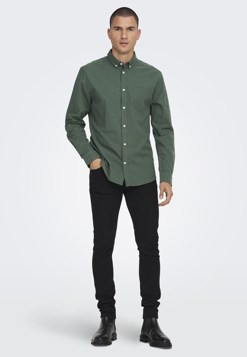 Camicia button-up verde scuro con colletto, dotata di taschino sul petto. Indossata con pantaloni neri slim-fit e stivaletti neri.