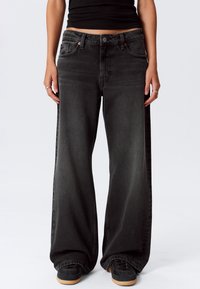 Persona che indossa jeans neri larghi, un top nero corto e sneakers nere con suole in gomma, in piedi contro uno sfondo bianco uniforme.
