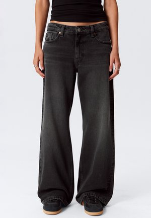 Persoon in losse zwarte denim spijkerbroek, zwart crop top, zwarte sneakers met gumzolen, staand tegen een effen witte achtergrond.