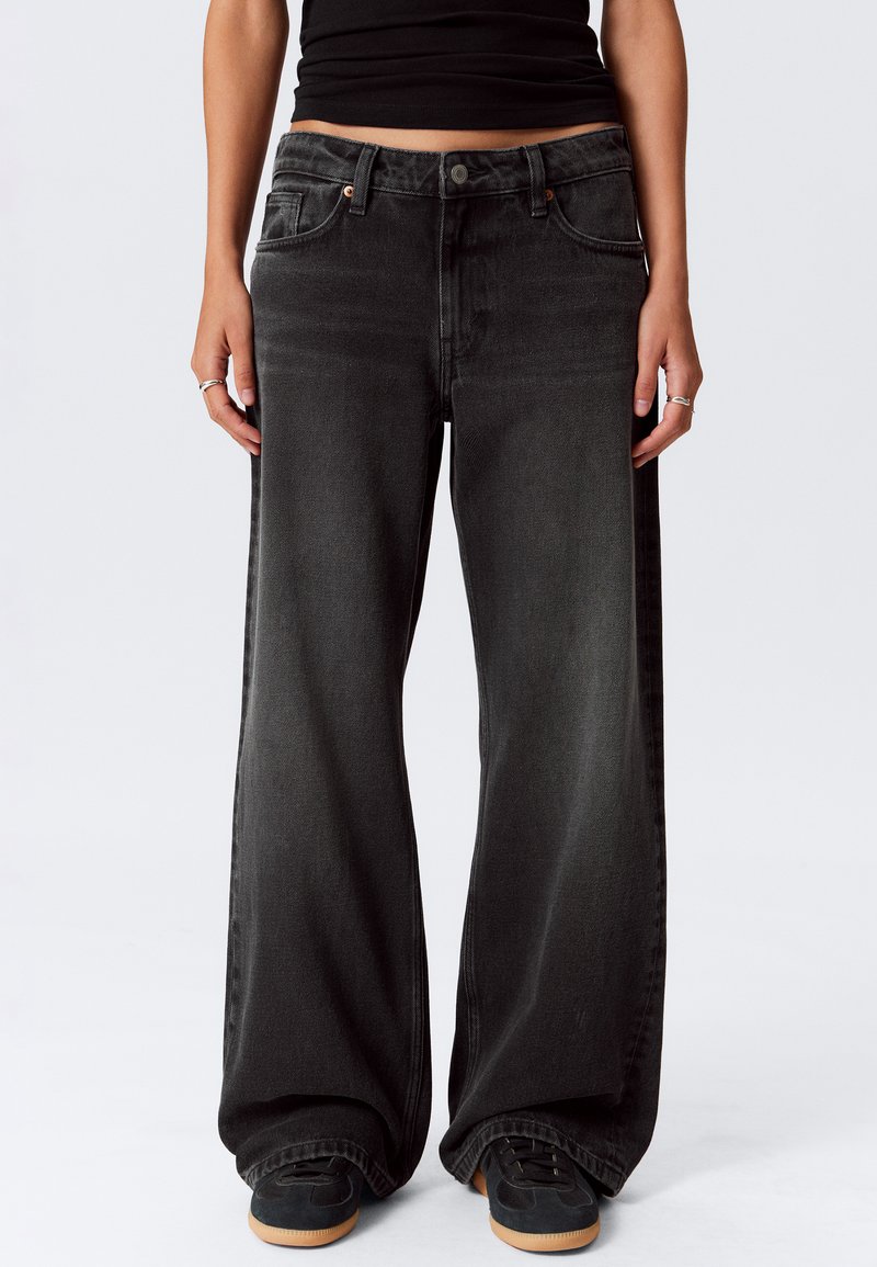 Persona che indossa jeans neri larghi, un top nero corto e sneakers nere con suole in gomma, in piedi contro uno sfondo bianco uniforme.