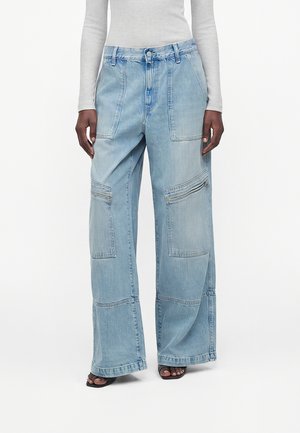 Světle modré džíny s širokými nohavicemi, velkými předními našitými kapsami a detaily na zip, nošené s bílým přiléhavým tričkem s dlouhým rukávem a černými sandály s otevřenou špičkou.