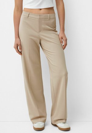 Frau trägt beige Hosen mit weitem Bein und weiße Sneakers, steht vor einem einfarbigen hellen Hintergrund.
