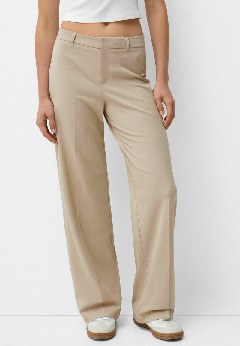 Femme portant un pantalon beige à jambes larges et des baskets blanches, debout devant un fond clair uni.