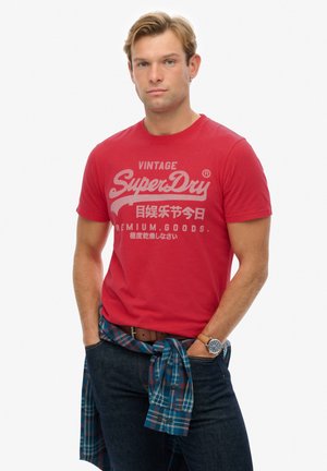 Red cotton t-shirt with white graphic print featuring "Vintage SuperDry" text. Worn with blue plaid shirt tied around waist and dark jeans.
