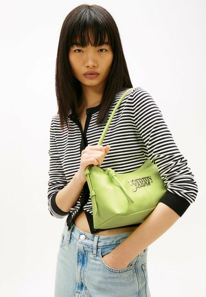 Jonge vrouw houdt limegroene Tommy Jeans handtas vast, draagt een zwart-wit gestreept shirt met lange mouwen en lichtblauwe spijkerbroek, kijkt vooruit.