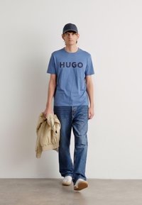 Mėlynas marškinėlių su apvaliu kaklu, ant kurio juodai spausdinta "HUGO", derinami su šviesiai mėlynomis džinsais. Modelis laiko bežį striukę ir vilkėjai baltus sportbačius.