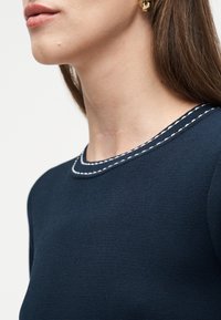 Κοντινό πλάνο του λαιμού και του κάτω μέρους του προσώπου ενός ατόμου που φοράει μπλε πουκάμισο ναυτικού με άσπρες ραφές και μαύρο σκουλαρίκι με χρυσή λεπτομέρεια.