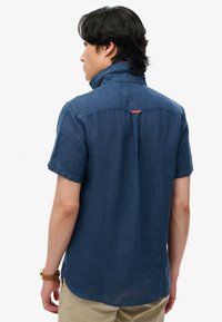 Homme aux cheveux foncés portant une chemise bleu marine à col relevé et manches courtes, ainsi qu'un pantalon beige, vu de dos sur un fond blanc.