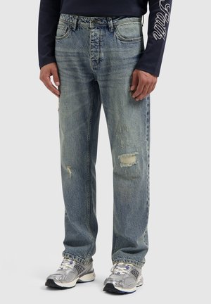 Lichtblauwe, versleten denim jeans met zichtbare scheuren, een rechte pasvorm en een licht vervaagde textuur. Draagt met grijze sportschoenen.