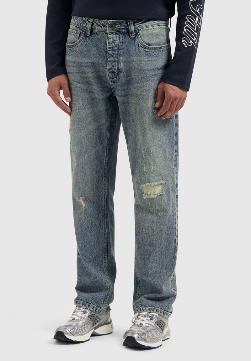 Lichtblauwe, versleten denim jeans met zichtbare scheuren, een rechte pasvorm en een licht vervaagde textuur. Draagt met grijze sportschoenen.
