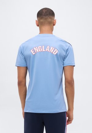 Bărbat cu păr scurt stă cu fața spre spate purtând o cămașă albastru deschis cu „ENGLAND” scris cu litere albe și roșii pe partea superioară a spatelui.