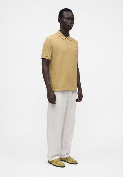 Un homme se tient debout, portant un polo beige, un pantalon blanc ample et des mocassins beiges sur fond blanc uni.