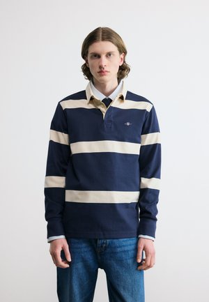 Junger Mann mit schulterlangem Haar, der ein marineblau-beige gestreiftes Rugbyhemd über einem Hemd mit Kragen und Krawatte trägt, vor einem schlichten weißen Hintergrund stehend.