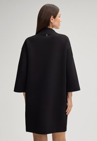 Zwart oversized jurkje met brede mouwen, gemaakt van een gladde, gestructureerde stof. Heeft een staande kraag en een klein logo accent aan de achterkant.