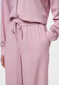 Blouse rose clair, ample avec fermeture à boutons et manches longues ; pantalons assortis à taille élastique avec cordon de serrage et poches latérales.
