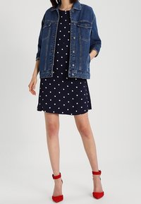 Námornická šaty s puntíky v kombinaci s džínovou bundou a červenými lodičkami na vysokém podpatku. Oblečení má klasický límec a dvě náprsní kapsy na bundě.