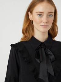 Ung kvinne med lys hud og fregner, iført svart bluse med volangdetaljer og sløyfe i sateng, gull hoop-øredobber, nøytral bakgrunn.