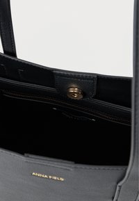 Čierna kožená taška typu tote s hladkou textúrou, mosadzným kovaním a zlatým logom vo vnútri. Obsahuje zapínanie na cvok a vrecko na zips.