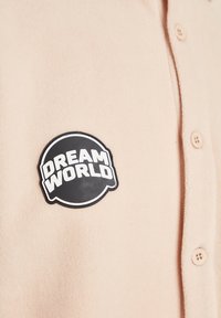 Chemise beige à boutons avec un patch circulaire noir sur la poitrine portant l'inscription "DREAM WORLD" en lettres blanches audacieuses.