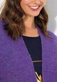 Lila texturerad kardiagan med v-ringning, med ett ribbat mönster. Bärs över en mörk topp med ett guld grafiskt accent.