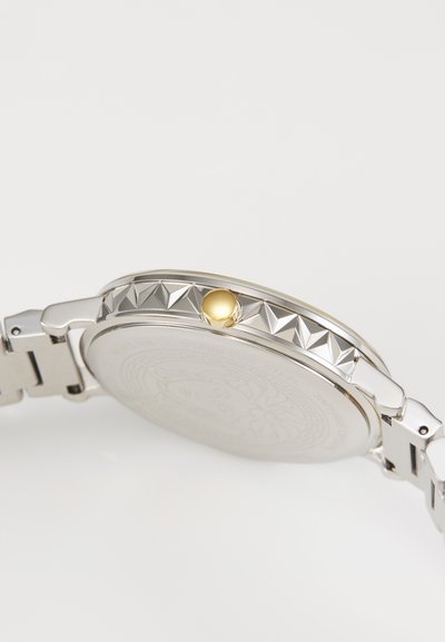 Montre en acier inoxydable argenté avec une lunette texturée et une couronne dorée. Présente un fond de boîtier lisse avec un design gravé et un bracelet.
