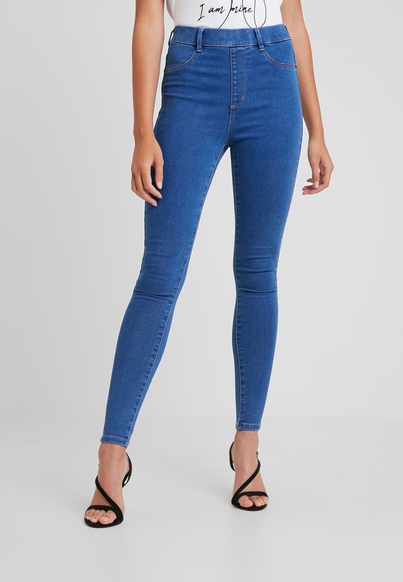 Femme portant un jean slim bleu et des sandales noires à talons hauts avec des lanières, debout devant un fond gris uni.