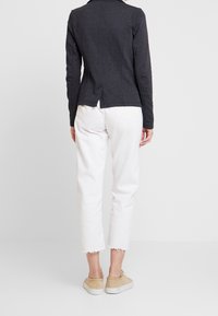 Chaqueta gris oscuro a medida con cuello, mangas largas y abertura trasera; combinada con vaqueros blancos recortados y zapatos beige.