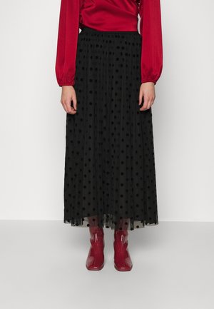 Kobieta nosząca czarną prześwitującą spódnicę midi w czarne kropki, czerwoną bluzkę z długim rękawem oraz błyszczące czerwone botki, stojąca na białym tle.