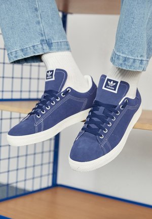 Námornícky modré tenisky Adidas vyrobené z semišu, s bielymi gumovými podrážkami, okrúhlymi špičkami a logom na jazyku. Viditeľné jasne biele ponožky.