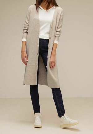 Vrouw draagt een lange beige cardigan met knopen over een wit shirt, donkere strakke jeans en witte sneakers, staand tegen een effen achtergrond.