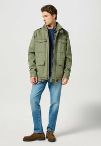 Olivgrön militärjacka i stil med fyra framfickor, ståkrage och avslappnad passform, bärs över en rutig skjorta och blå jeans.