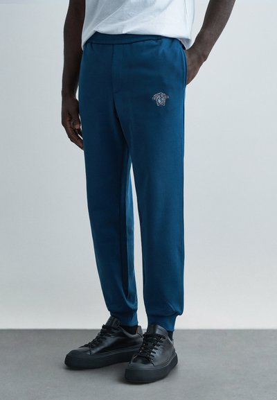 Blue sweatpants with a slim fit, elastic waistband, side pockets, and embroidered logo detail. Paired with black sneakers.