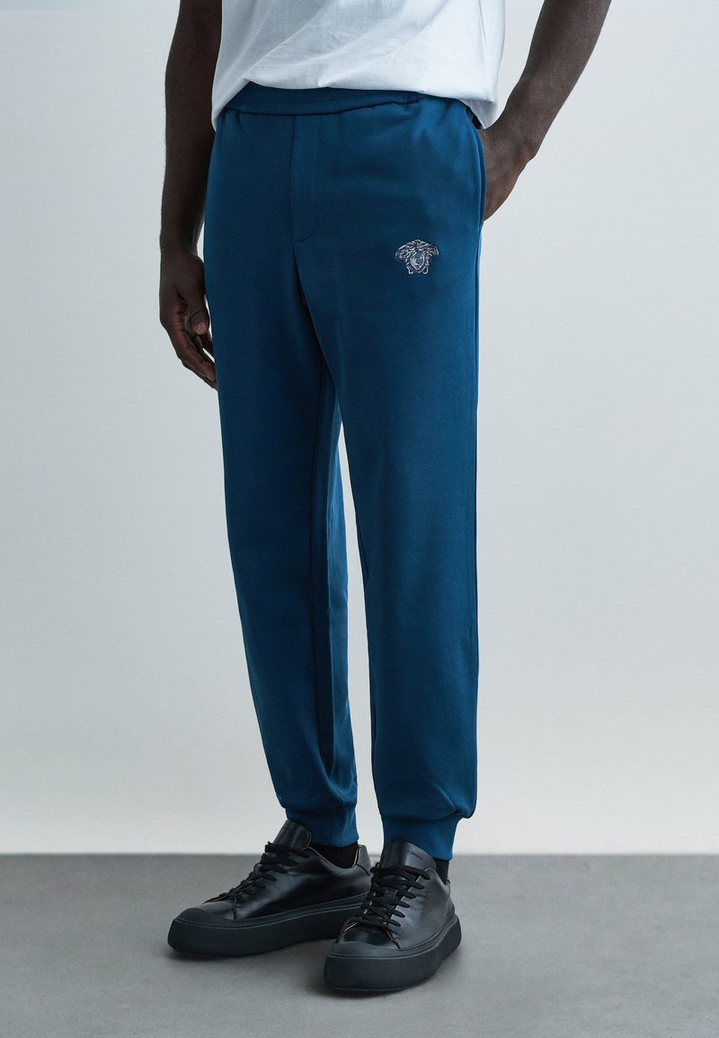 Modré tepláky so slim strihom, elastickým pásom, bočnými vreckami a vyšívaným logom. Kombinované s čiernymi teniskami.