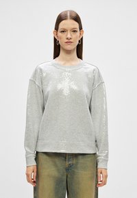 Mulher com cabelo castanho liso, vestindo um suéter prateado com lantejoulas e calças largas de um verde desbotado, de pé contra um fundo simples.