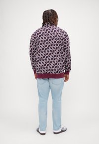 Veste en maille dans un motif géométrique violet, crème et rouge. Dotée d'un col et de poignets en côte bordeaux. Portée avec un jean bleu clair et des chaussures à rayures noires.