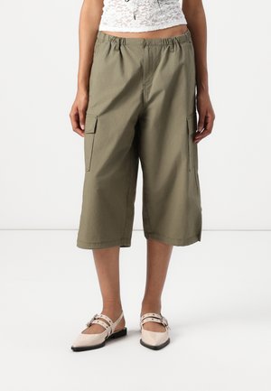 Persona que lleva pantalones cortos de pierna ancha de color verde oliva con bolsillos laterales y bailarinas puntiagudas beige con doble correa.