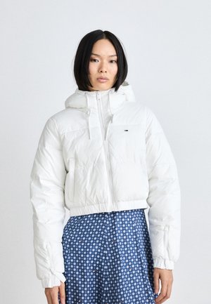 Chaqueta blanca pompón con capucha, con cierre de cremallera y puños elásticos. Lleva puesta sobre un vestido azul con estampado. Textura suave, diseño corto.